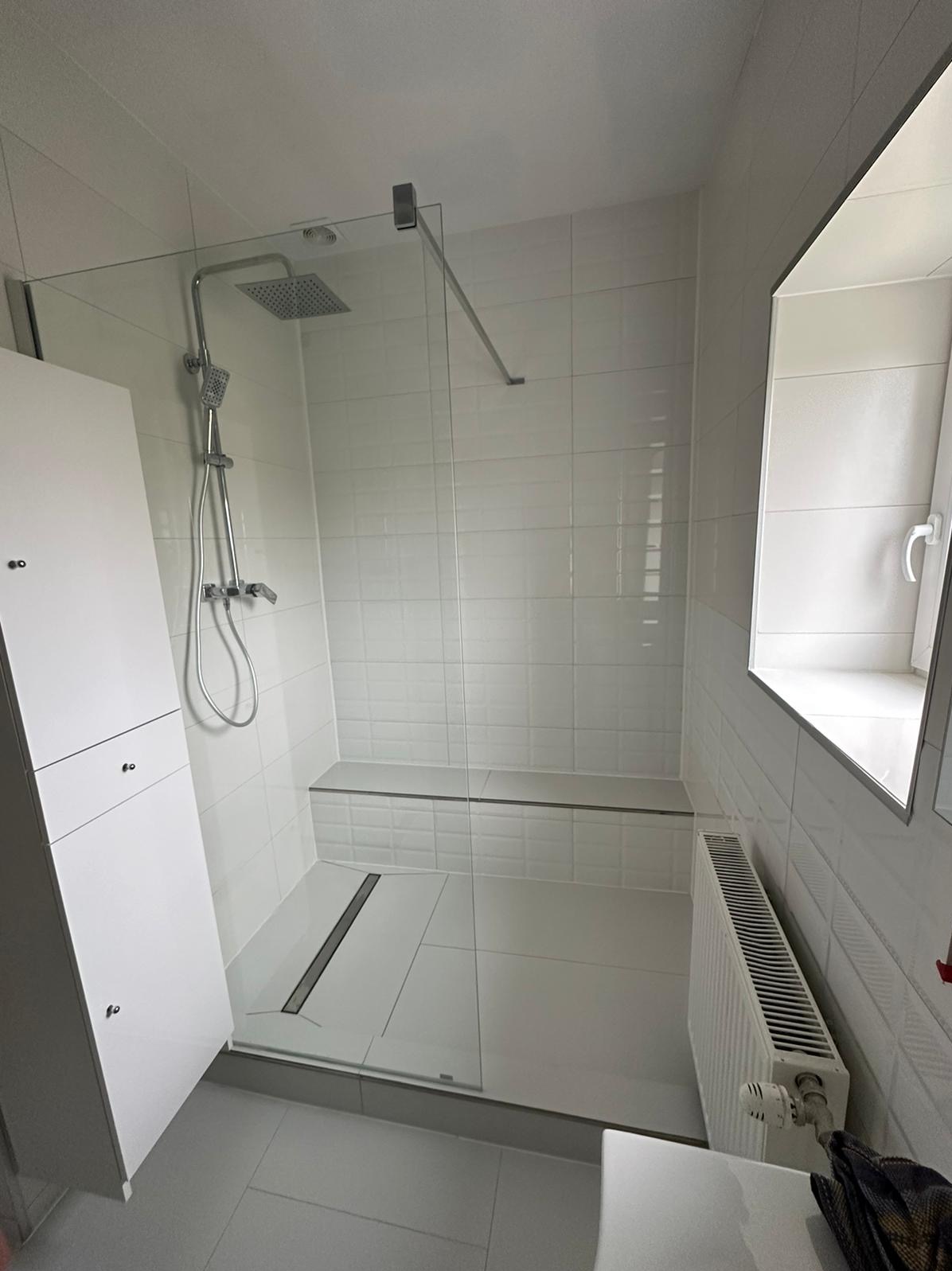 Nowoczesna kabina prysznicowa walk-in z odpływem liniowym, wykończona białymi płytkami, z siedziskiem i grzejnikiem pod oknem.