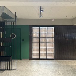 S.A.R.O - Nowoczesne wykończenie wnętrza z ciemną, prążkowaną okładziną ścienną, zielonymi drzwiami i metalową konstrukcją z półkami na rośliny, oświetlona witryna w tle.