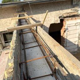 S.A.R.O - Zbrojenie balkonu z drewnianym szalunkiem i pianką montażową, widok z góry na konstrukcję na tle budynku z oknem i otworem w murze, w słoneczny dzień.