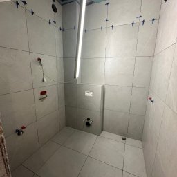 S.A.R.O - Wnętrze łazienki w trakcie wykańczania: szare płytki na ścianach i podłodze, niebieskie krzyżyki dystansowe, widoczne instalacje. Oświetlenie robocze.