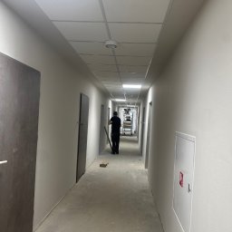 S.A.R.O - Długi, jasny korytarz w trakcie malowania. Widoczny pracownik z narzędziami, drabina w oddali i świeżo pomalowane ściany w odcieniach bieli i szarości.