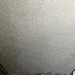 S.A.R.O - Szara, teksturowana powierzchnia sufitu z bliska, ukazująca nieregularny wzór i fakturę. Widoczny fragment podłogi i wiadro z farbą w rogu kadru.