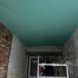 S.A.R.O - Montaż zielonych płyt gipsowych na suficie w surowym wnętrzu. Widoczne rusztowanie, cegła i pustaki. Etap prac wykończeniowych.