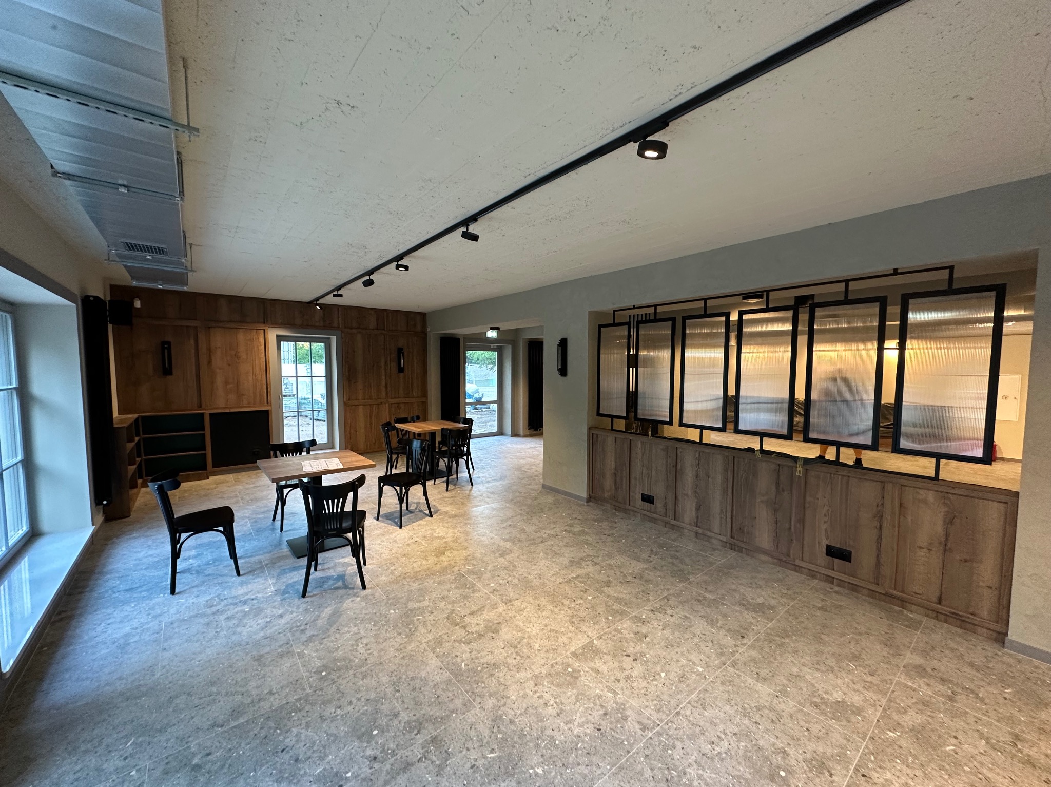 Przestronne wnętrze po remoncie: szara, wzorzysta podłoga, drewniane panele ścienne, czarne krzesła i stoły. Widoczne okna i oświetlenie sufitowe.