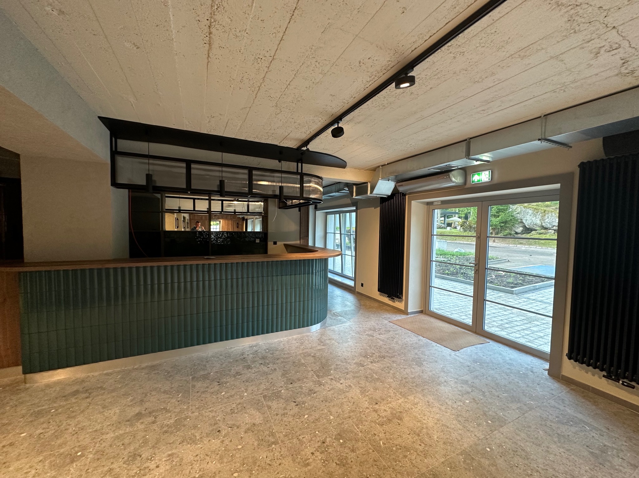 Nowoczesne wnętrze z zieloną ladą recepcyjną z płytek, szarym kamiennym podłożem i surowym betonowym sufitem z oświetleniem punktowym. Duże okna wpuszczają naturalne światło.