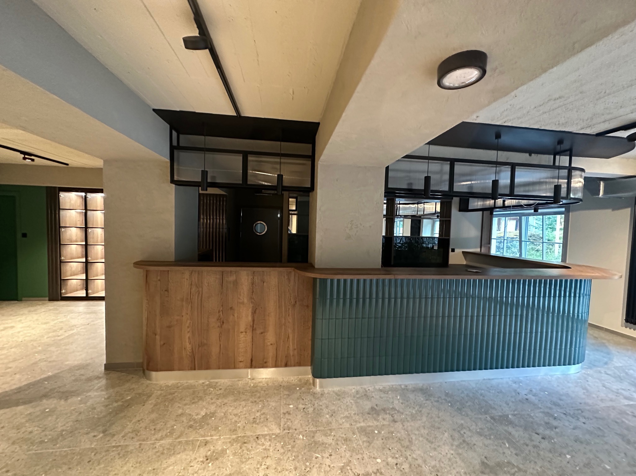 Wykończona przestrzeń komercyjna z drewnianą ladą i ścianą z zielonych, pionowych płytek. Widoczne oświetlenie sufitowe i podłoga z lastryko.