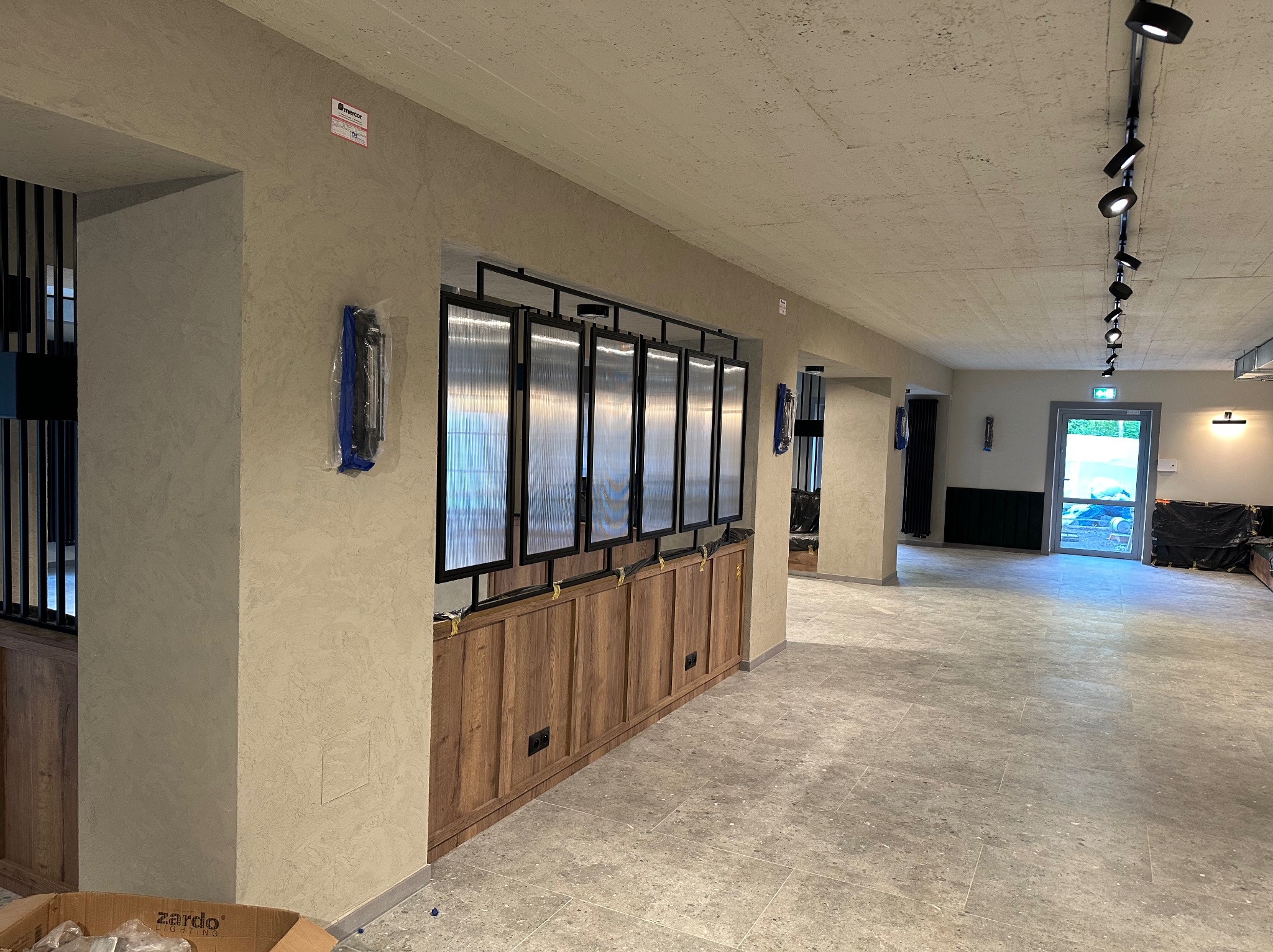 Wnętrze z wykończoną ścianą w odcieniach beżu, drewnianą zabudową z ryflowanymi szybami i szarą posadzką. Oświetlenie punktowe na suficie.