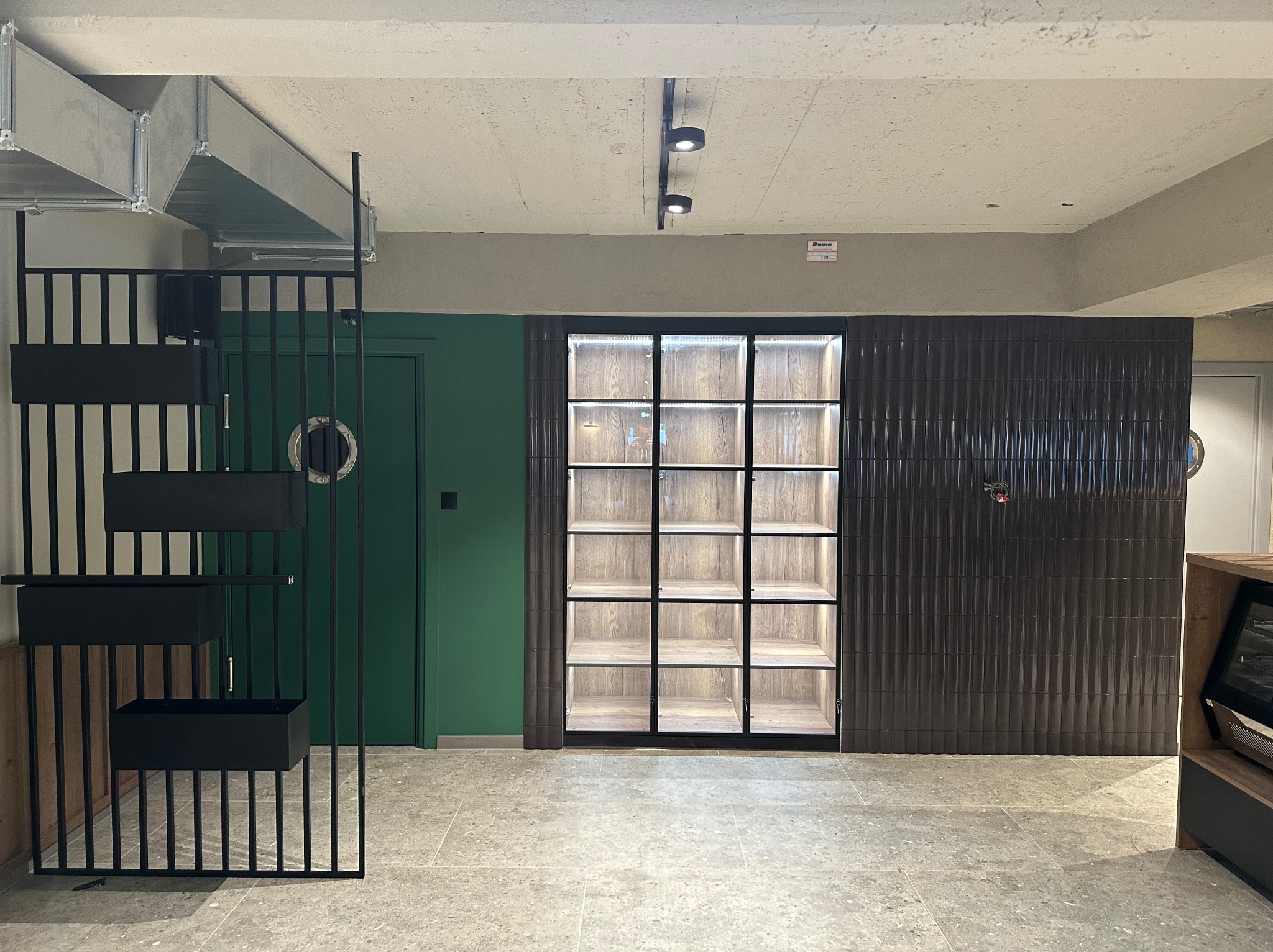 Nowoczesne wykończenie wnętrza z ciemną, prążkowaną okładziną ścienną, zielonymi drzwiami i metalową konstrukcją z półkami na rośliny, oświetlona witryna w tle.