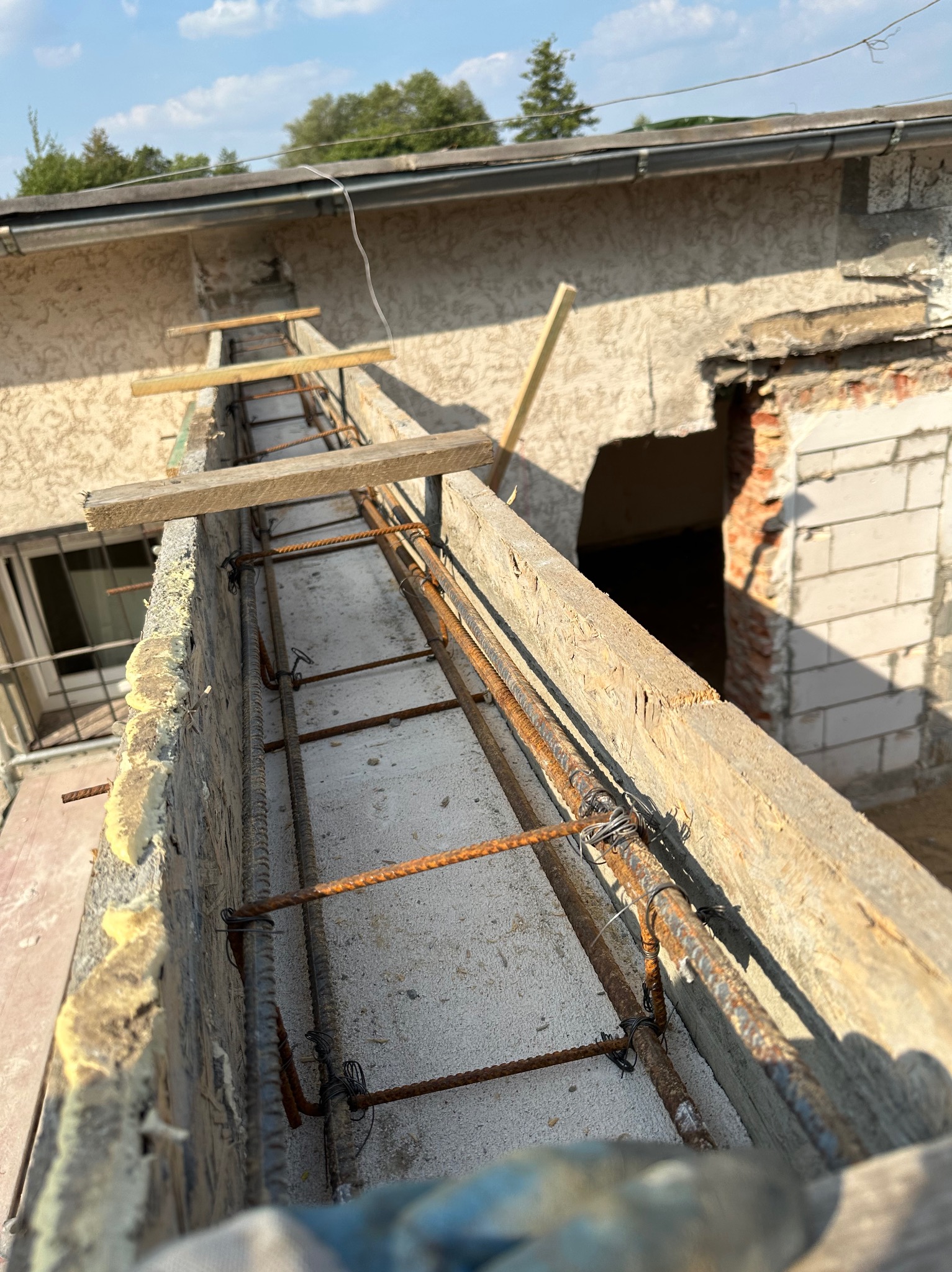 Zbrojenie balkonu z drewnianym szalunkiem i pianką montażową, widok z góry na konstrukcję na tle budynku z oknem i otworem w murze, w słoneczny dzień.