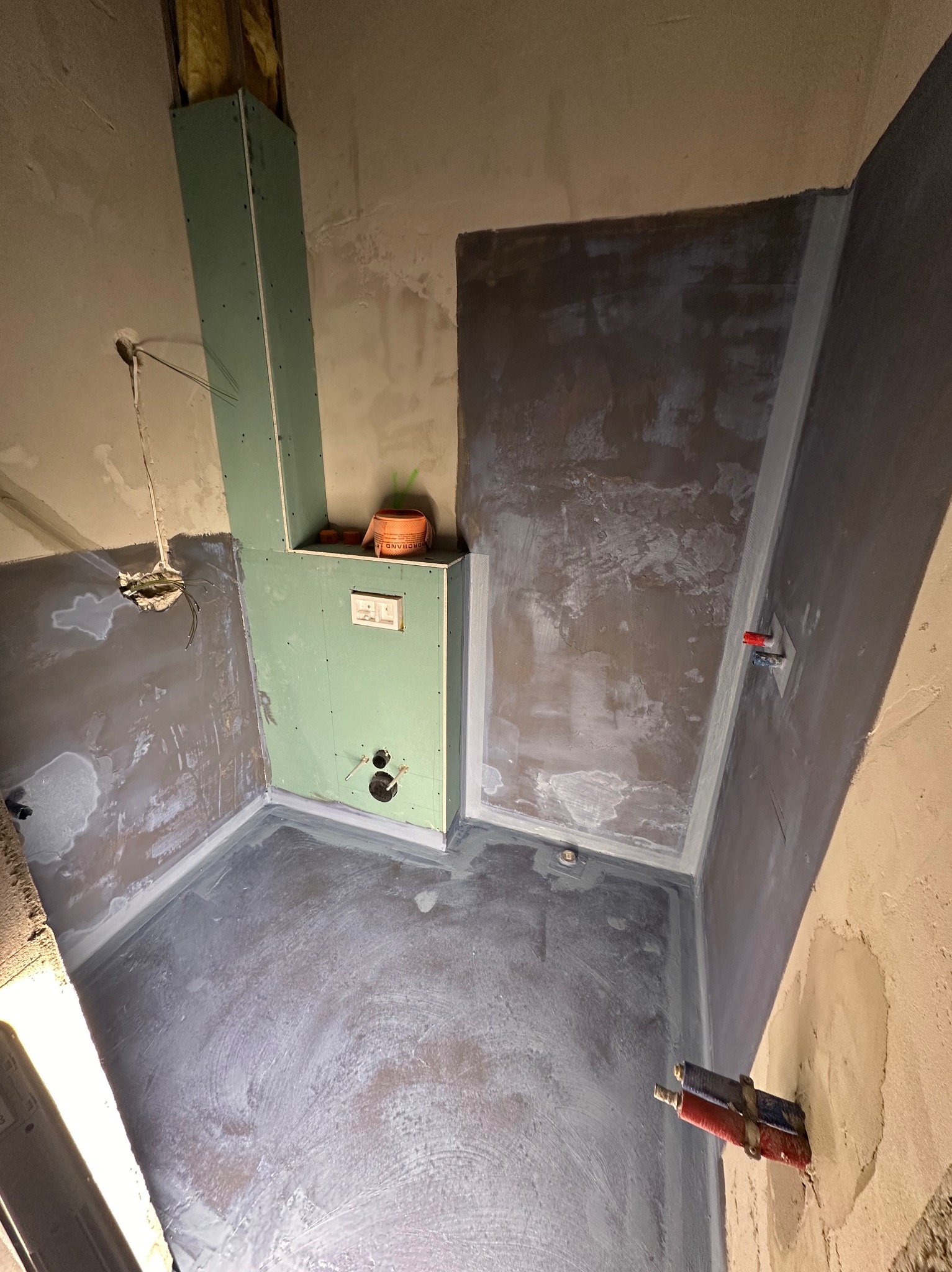 Łazienka w trakcie hydroizolacji: ściany i podłoga pokryte szarą warstwą, widoczne rury, zabudowa z płyt gipsowych. Etap prac przed wykończeniem.