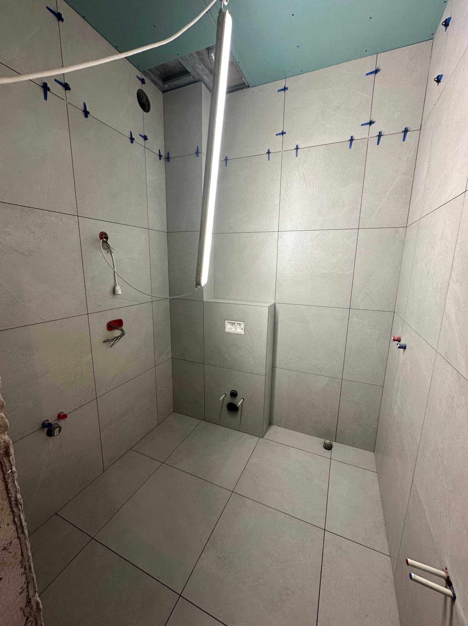 Wnętrze łazienki w trakcie wykańczania: szare płytki na ścianach i podłodze, niebieskie krzyżyki dystansowe, widoczne instalacje. Oświetlenie robocze.