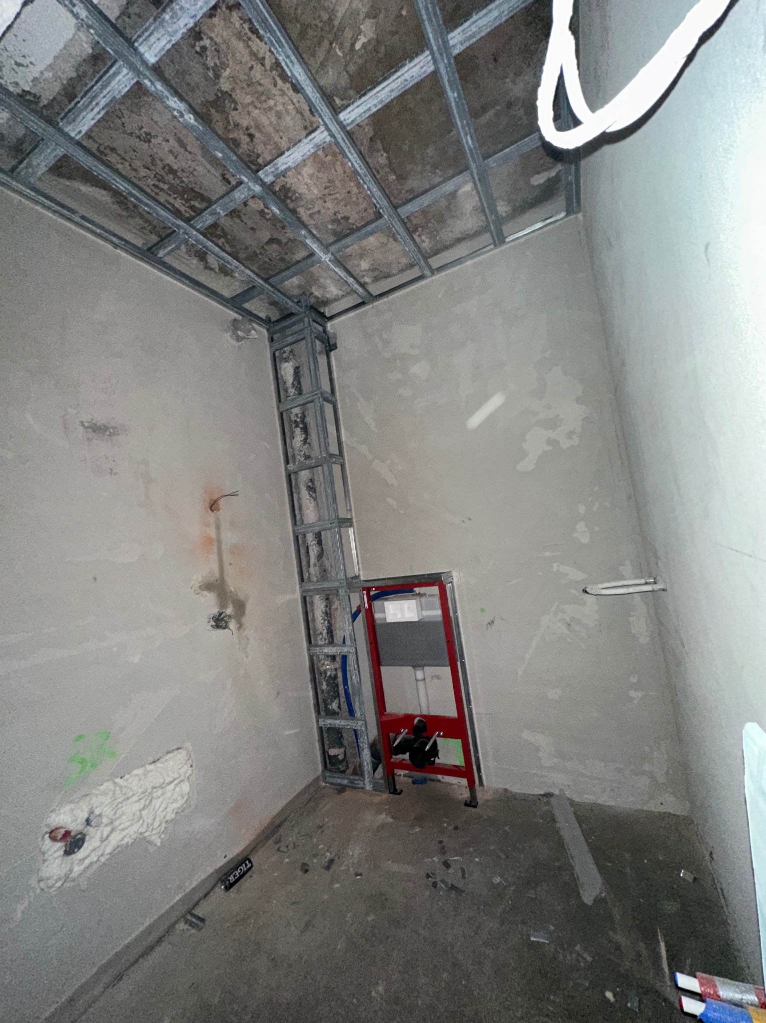 Wnętrze łazienki w trakcie remontu: widoczne metalowe profile sufitu podwieszanego, stelaż podtynkowy WC z czerwonymi elementami i instalacje na ścianach.
