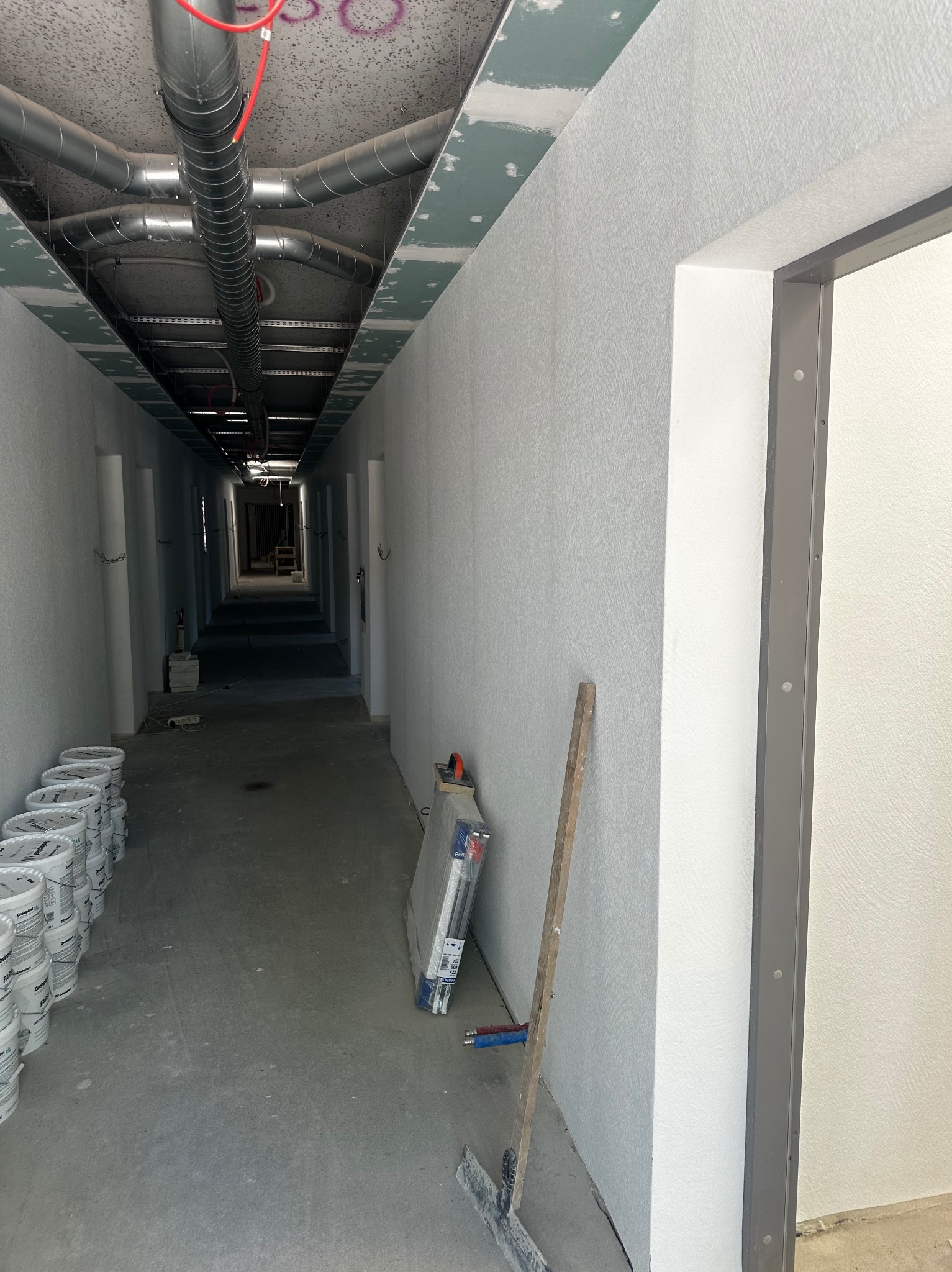 Korytarz w trakcie wykańczania: białe ściany z fakturą, widoczne narzędzia i puste wiadra po farbie. Sufit z instalacją wentylacyjną. Ościeżnica drzwiowa.