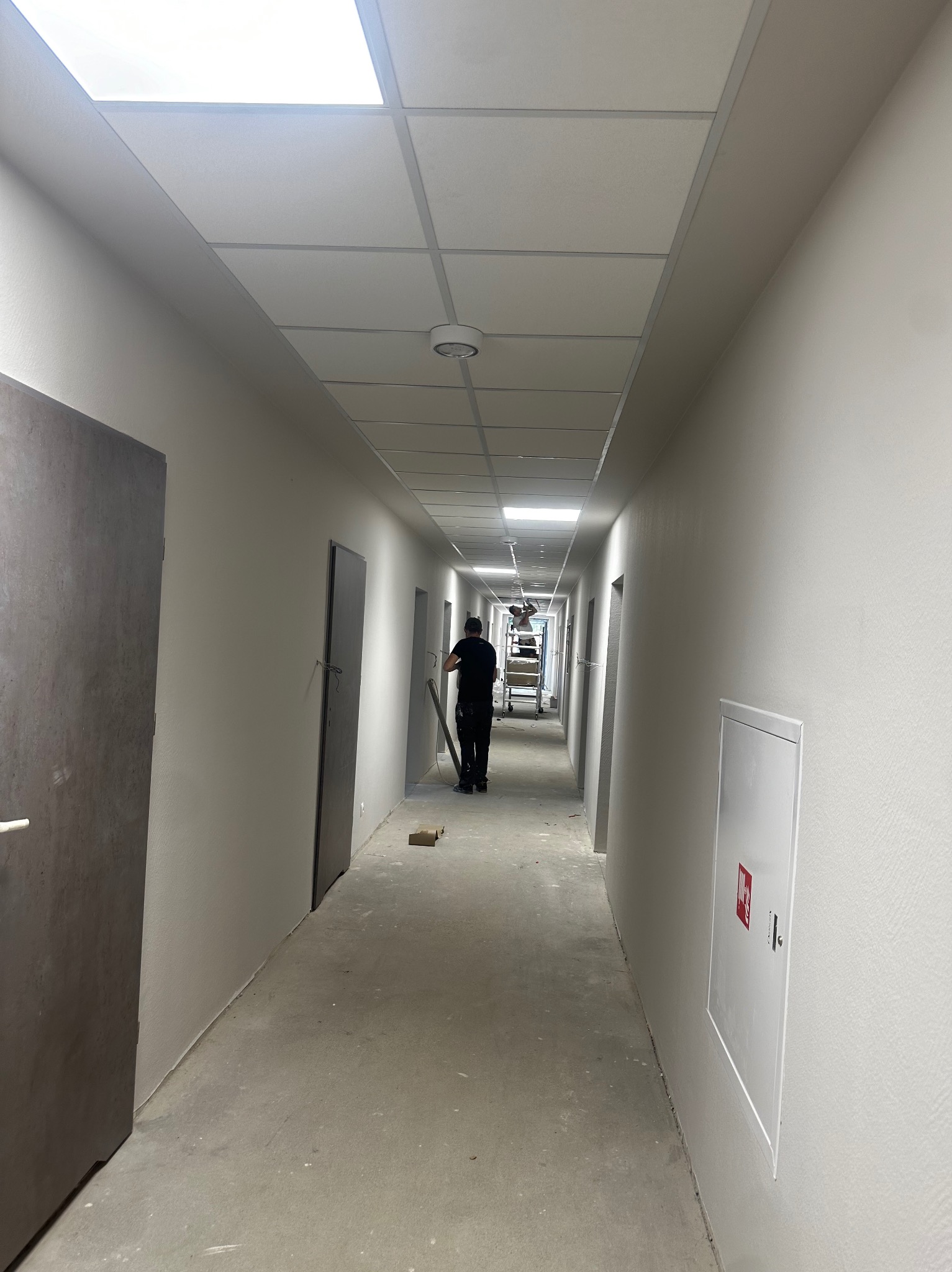 Długi, jasny korytarz w trakcie malowania. Widoczny pracownik z narzędziami, drabina w oddali i świeżo pomalowane ściany w odcieniach bieli i szarości.