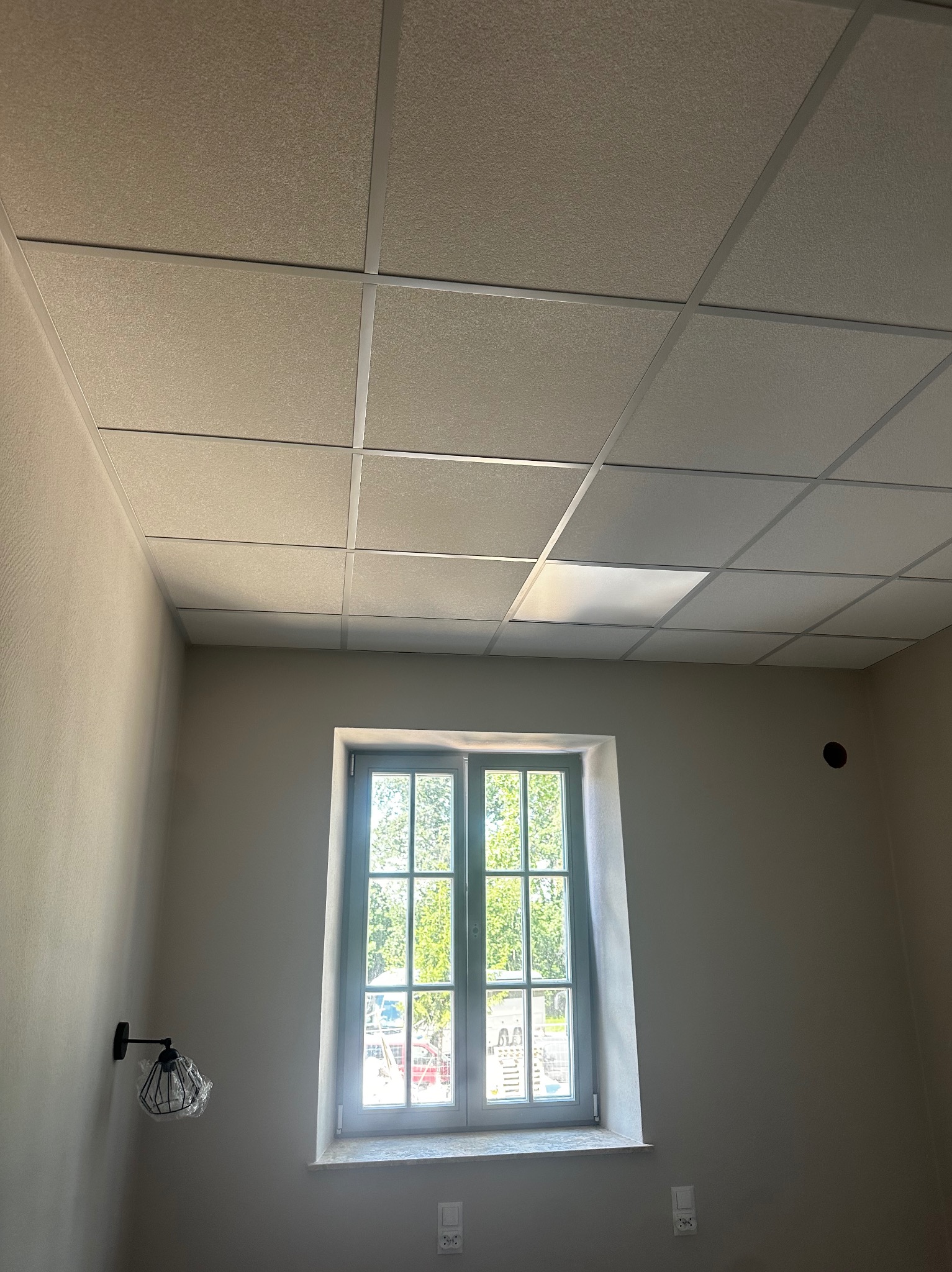 Kętrzyn: Pokój po remoncie z oknem i sufitem podwieszanym. Ściany pomalowane na szaro, widoczny nowy kinkiet i gniazdka elektryczne. Jasne wnętrze.
