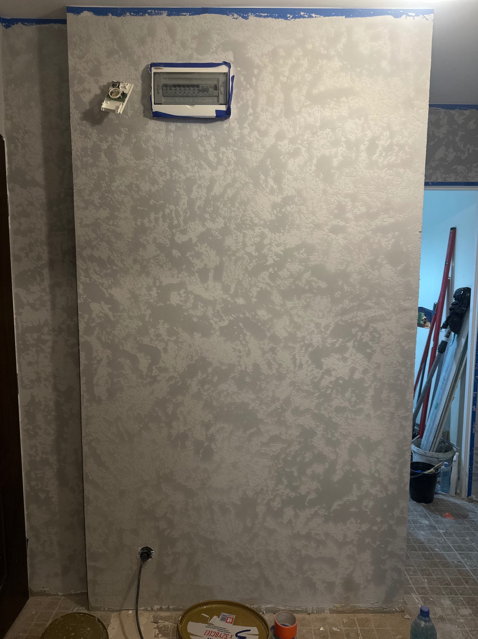 Ściana z dekoracyjną, szarą strukturą, zabezpieczona niebieską taśmą malarską przy suficie, z widoczną skrzynką elektryczną i puszką instalacyjną. W tle drabina i narzędzia.