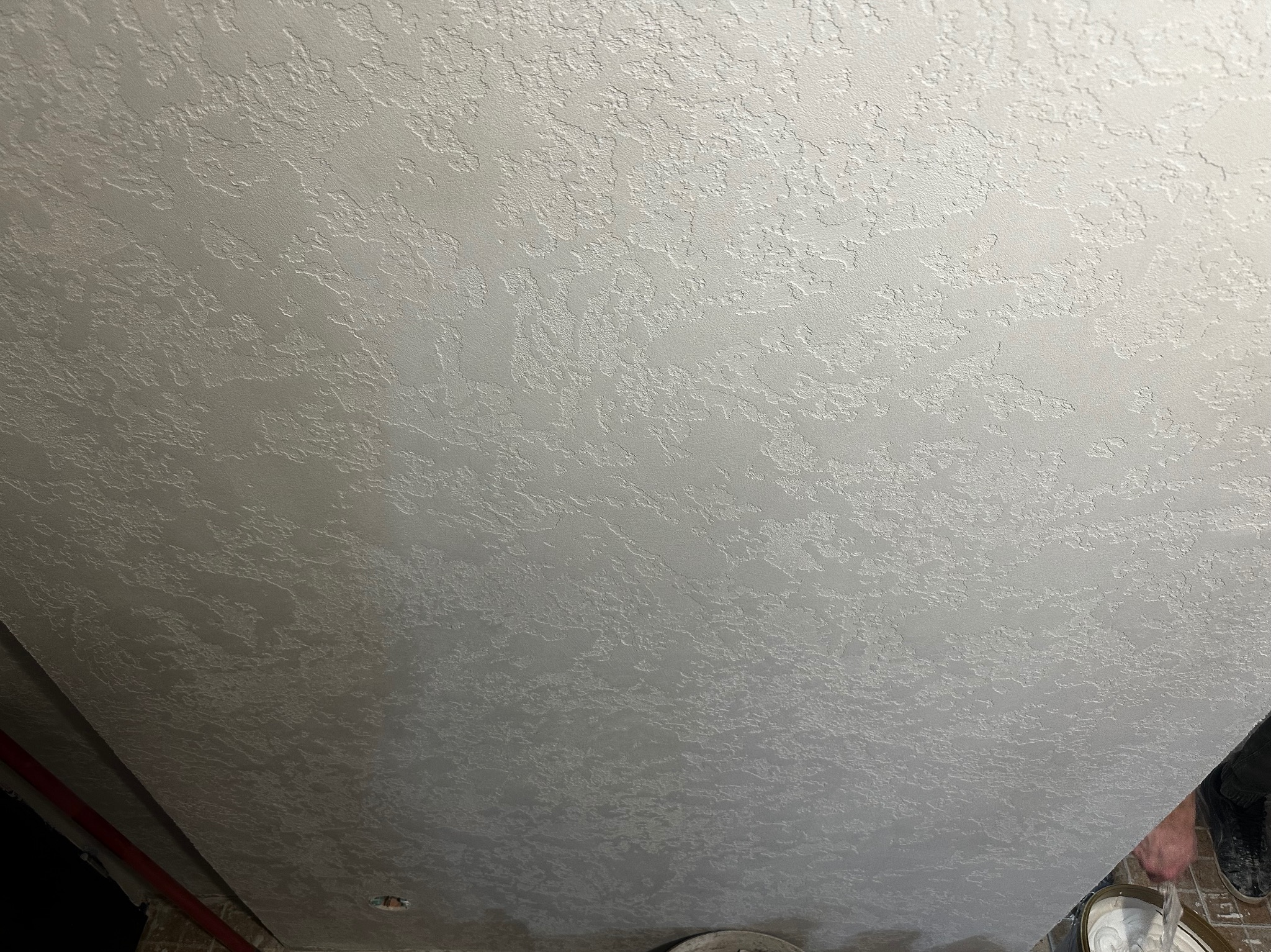 Szara, teksturowana powierzchnia sufitu z bliska, ukazująca nieregularny wzór i fakturę. Widoczny fragment podłogi i wiadro z farbą w rogu kadru.