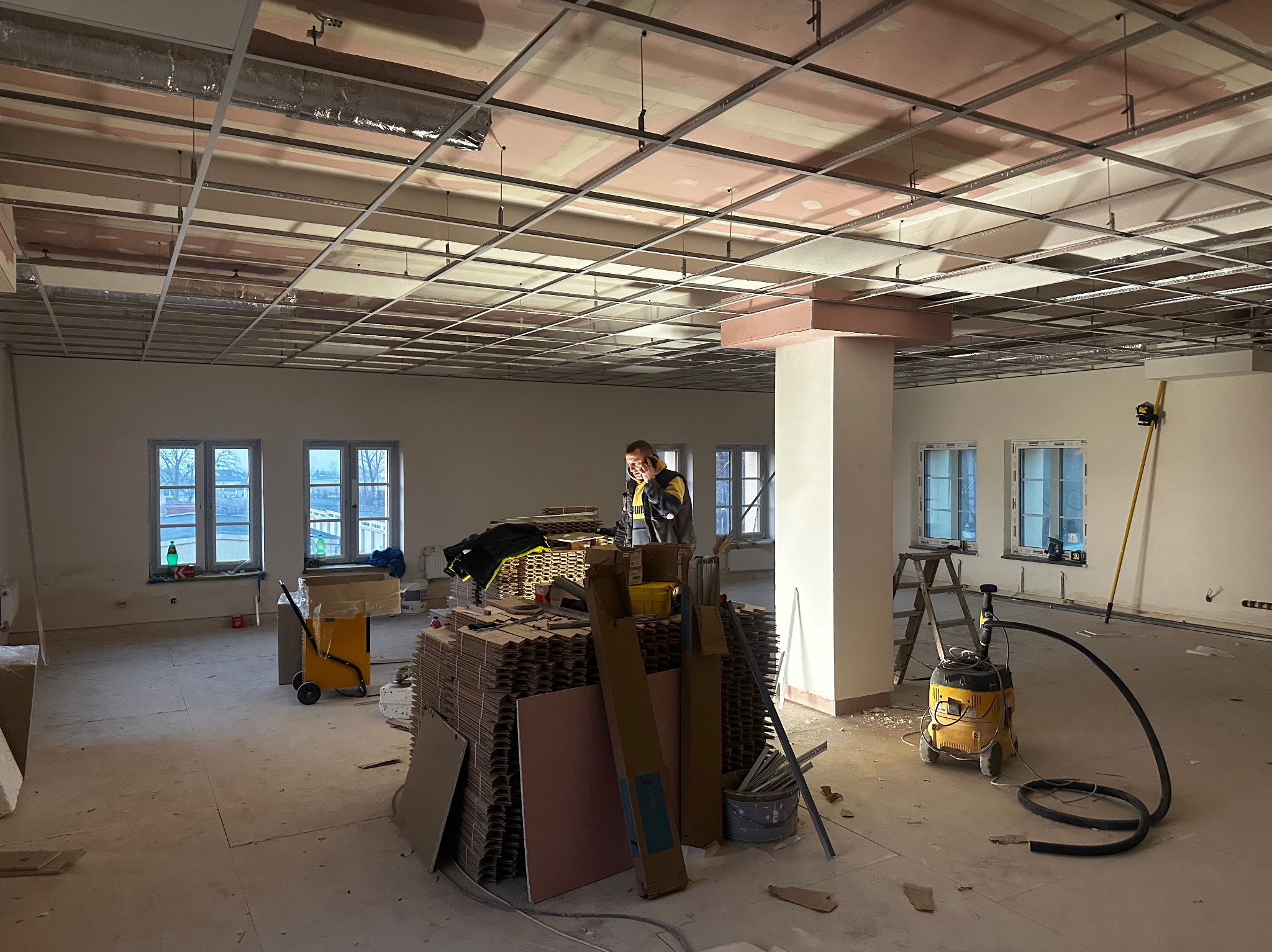 Montaż sufitu podwieszanego w przestronnym pomieszczeniu. Widoczne profile, płyty gipsowe i narzędzia. Pracownik rozmawia przez telefon. W tle okna.