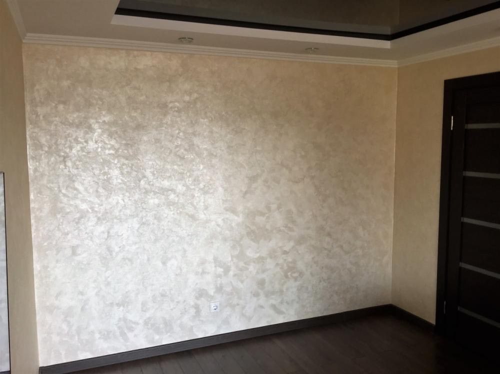 Ściana z dekoracyjną, perłową fakturą w odcieniach beżu i bieli, widoczna w rogu ciemna, nowoczesna stolarka drzwiowa i ciemna podłoga z paneli.