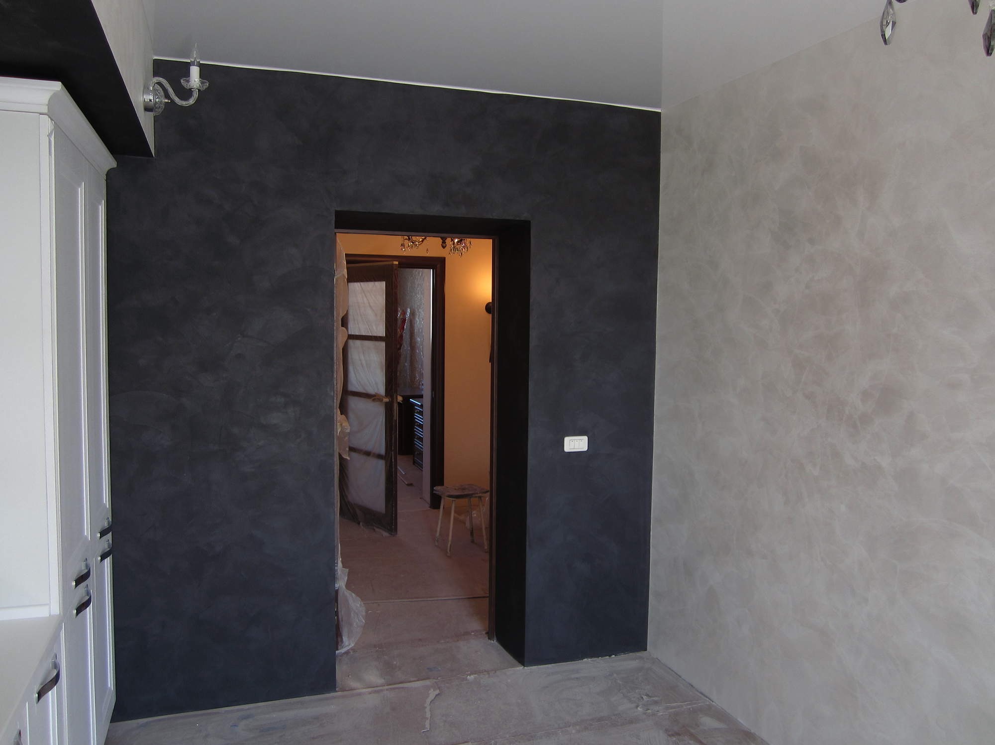Ściana z efektem dekoracyjnym w odcieniach szarości i grafitu, widoczna wnęka drzwiowa i fragment białej szafki. W tle widoczny pokój z żyrandolem.