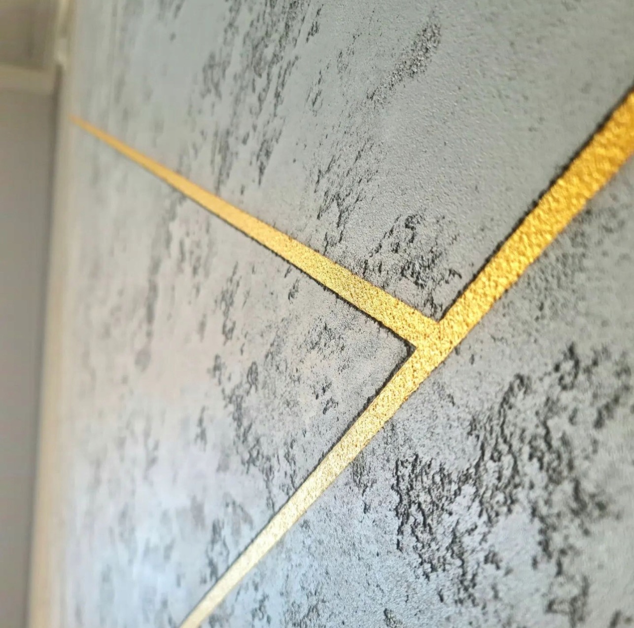 Dekoracyjna ściana z szarą fakturą i złotymi, ukośnymi liniami tworzącymi geometryczny wzór. Efektowna aranżacja wnętrza z bliska.