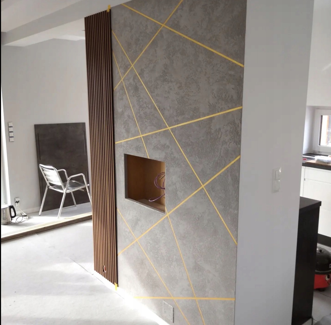 Ściana z dekoracyjną szarą strukturą, zdobiona żółtymi liniami i drewnianymi lamelami. W ścianie wnęka z widocznymi kablami. W tle widoczne krzesło i elementy wyposażenia.