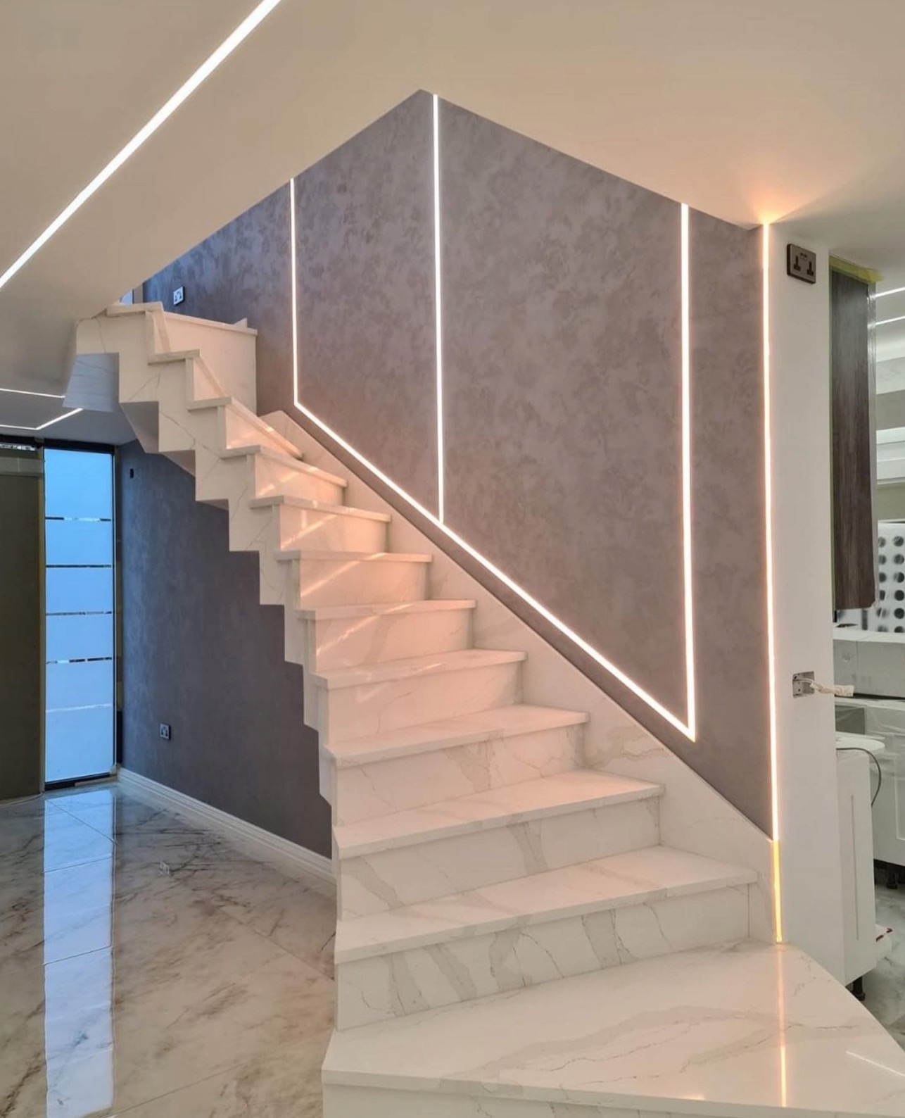 Nowoczesne schody z białego marmuru z podświetleniem LED na ścianie o fakturze betonu. Eleganckie wykończenie wnętrza z dbałością o detale i oświetlenie.