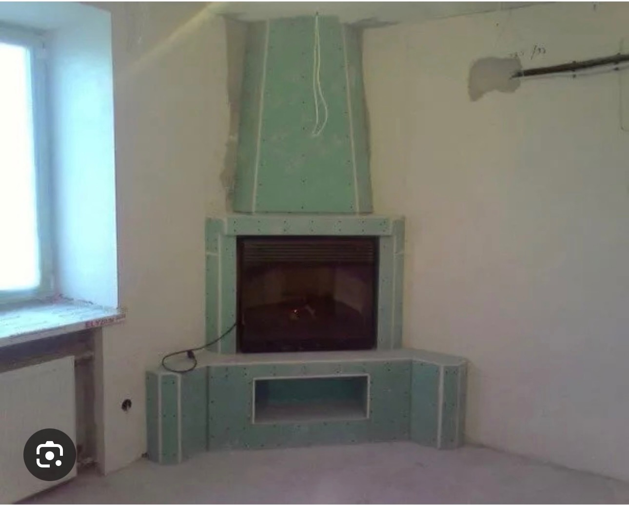 Obudowa kominka z płyt gipsowo-kartonowych w trakcie montażu, widok na surową konstrukcję przed wykończeniem, w tle okno i fragment ściany.