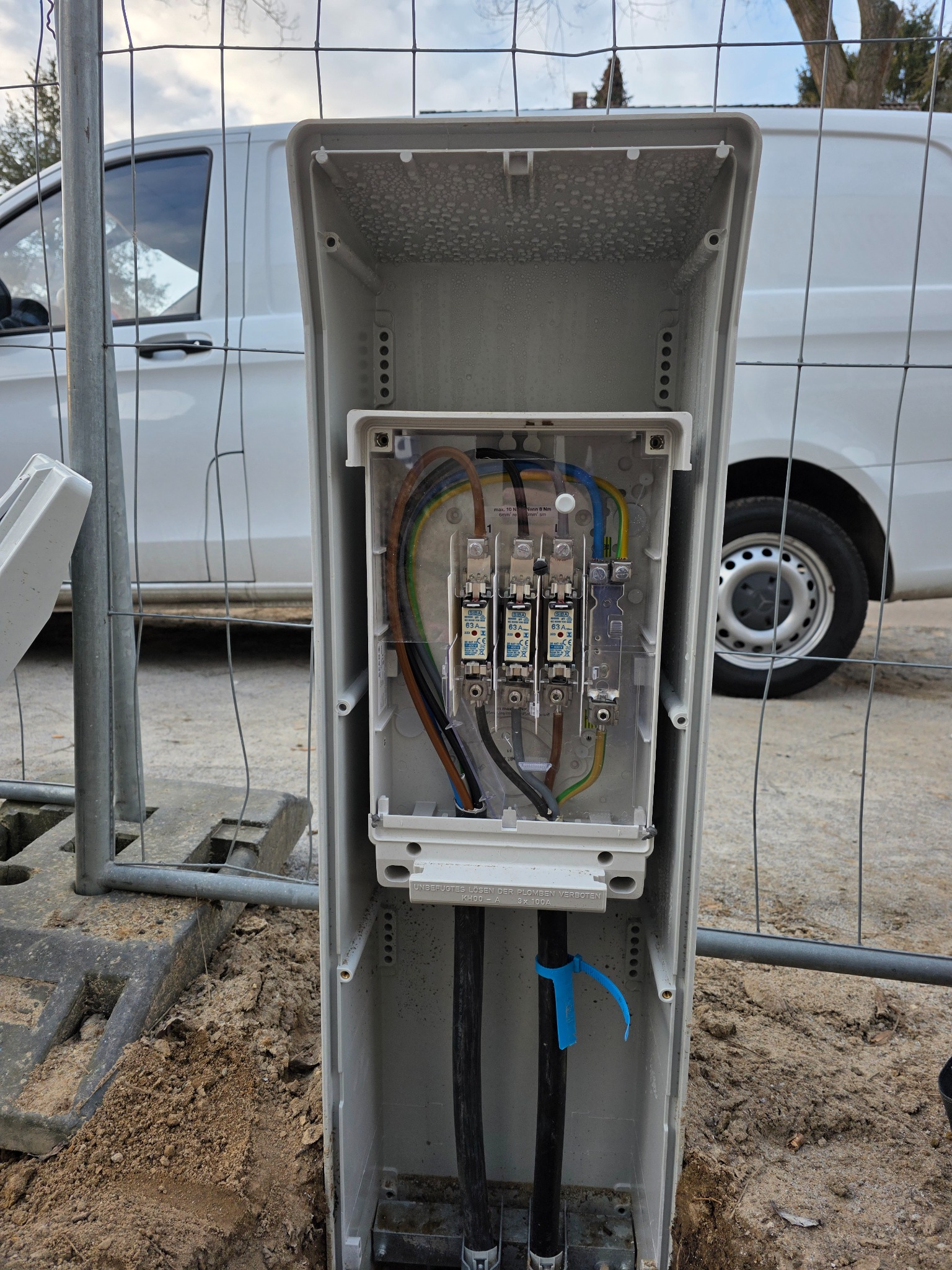 Otwarta skrzynka elektryczna z bezpiecznikami i okablowaniem, widoczna przez metalową siatkę ogrodzeniową, z białym samochodem dostawczym w tle.