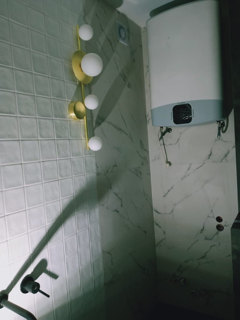 Wnętrze łazienki z białymi płytkami kwadratowymi, marmurowymi panelami, dekoracyjną złotą lampą z kulistymi kloszami i podgrzewaczem wody na ścianie.