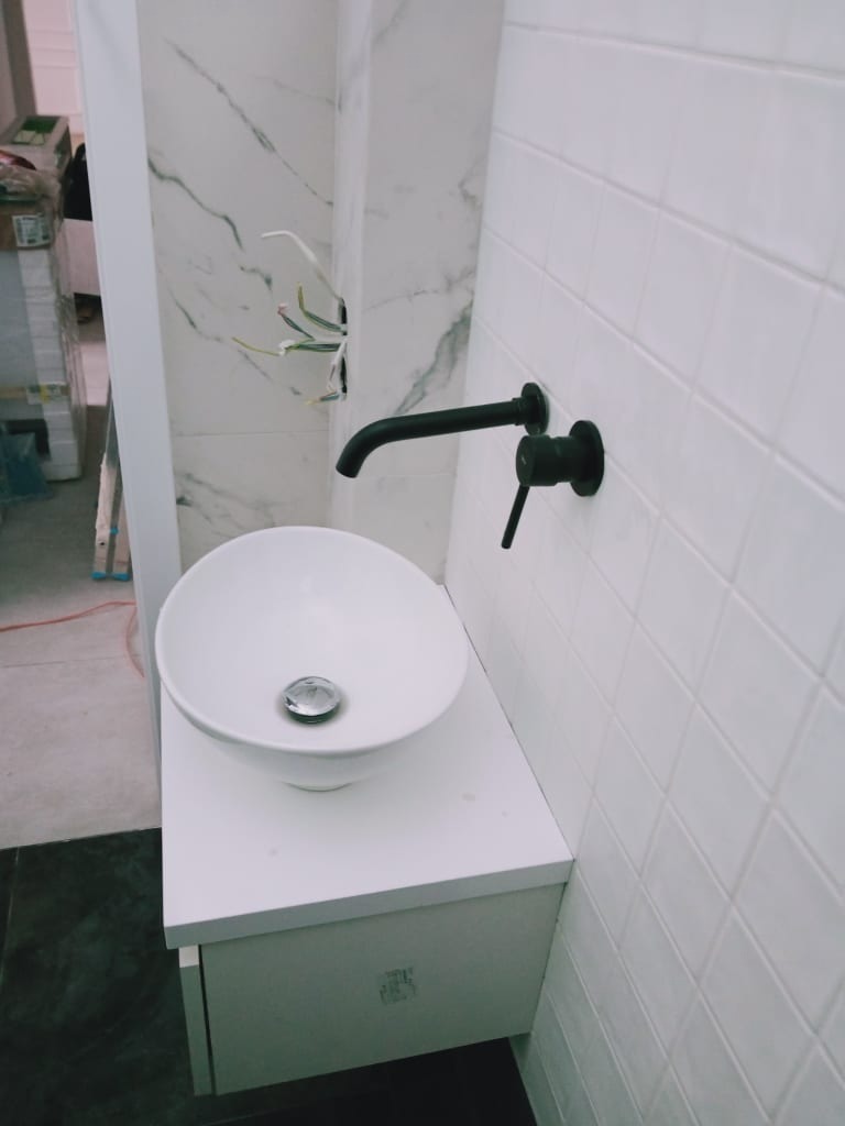 Biała umywalka nablatowa z czarną armaturą na szafce w łazience wykończonej białymi, kwadratowymi płytkami i marmurowymi panelami, widoczne wystające kable.