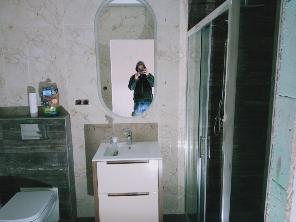Wnętrze łazienki w trakcie prac wykończeniowych: widoczna kabina prysznicowa, umywalka z szafką, toaleta i lustro owalne, w którym odbija się osoba robiąca zdjęcie telefonem.