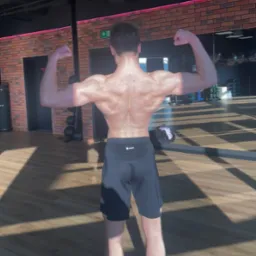 Mężczyzna prezentuje sylwetkę po treningu na siłowni, widok z tyłu, napięte mięśnie pleców i ramion, na podłodze widoczne cienie od promieni słonecznych.