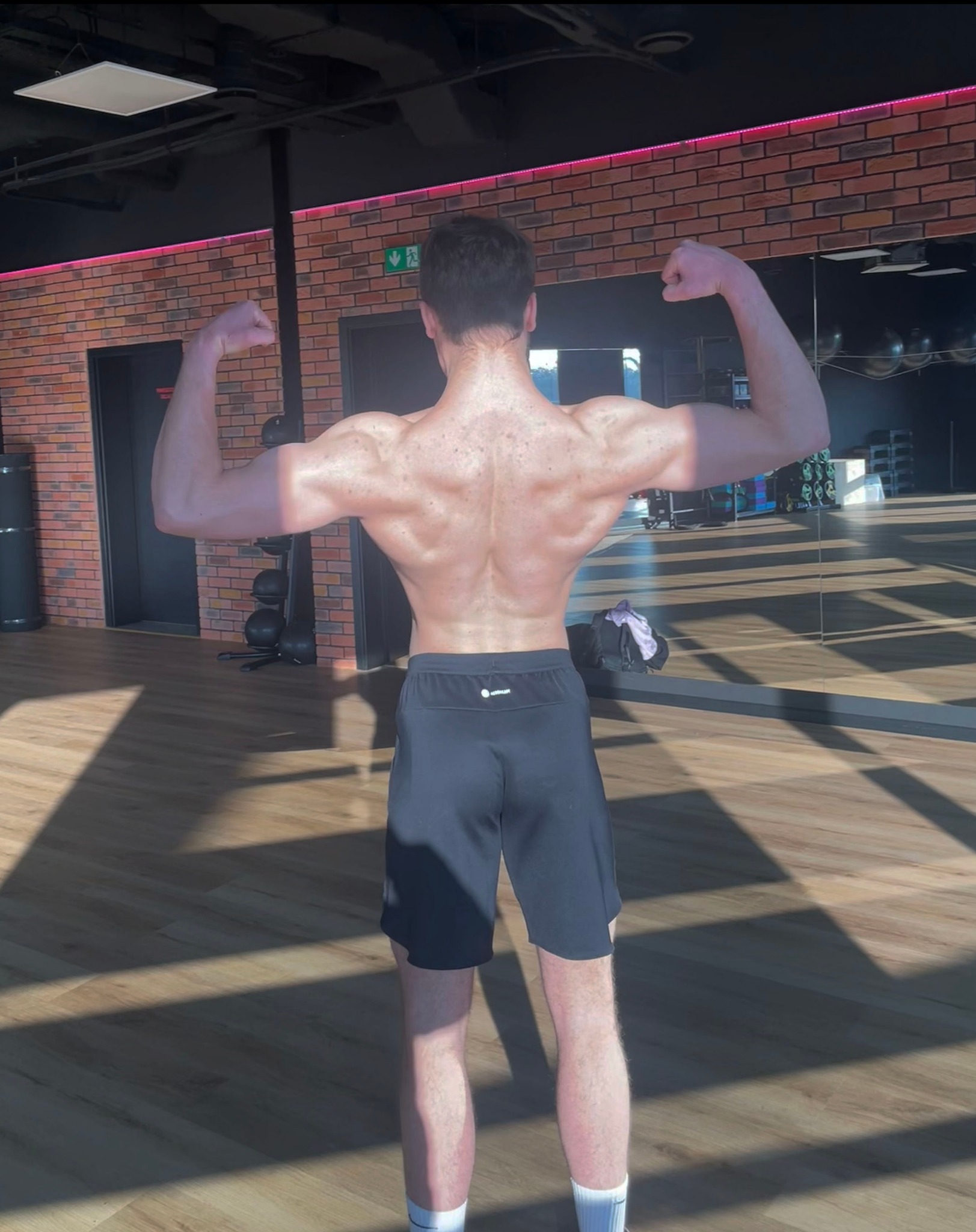 Mężczyzna prezentuje sylwetkę po treningu na siłowni, widok z tyłu, napięte mięśnie pleców i ramion, na podłodze widoczne cienie od promieni słonecznych.