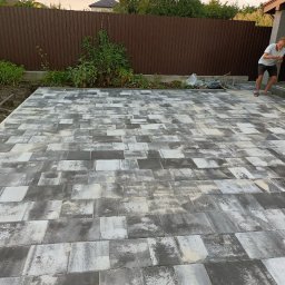 Artem Vynnychuk - Ułożona szara kostka brukowa na posesji, widoczny fragment brązowego ogrodzenia i roślinność przy granicy z kostką, w tle sylwetka mężczyzny.