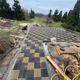 Artem Vynnychuk - Ułożona kostka brukowa w szachownicę z brązowych, czarnych i beżowych elementów, widoczny fragment obrzeża i narzędzia, tło z drzewami i pochmurnym niebem.