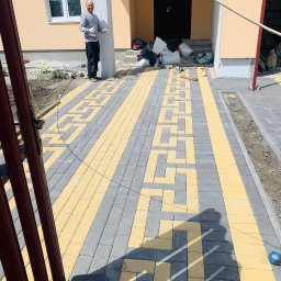 Artem Vynnychuk - Ułożona kostka brukowa w geometryczny wzór z żółtych i szarych elementów przed wejściem do domu; pracownik budowlany pozuje z uniesionymi kciukami, obok leżą worki z materiałami budowlanymi.