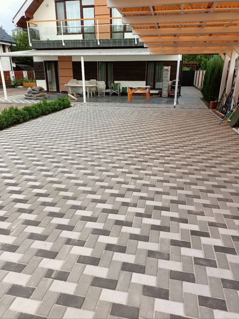 Nowo ułożony podjazd z kostki brukowej w odcieniach szarości, wzór jodełka, z widokiem na dom z balkonem i zadaszone miejsce parkingowe.
