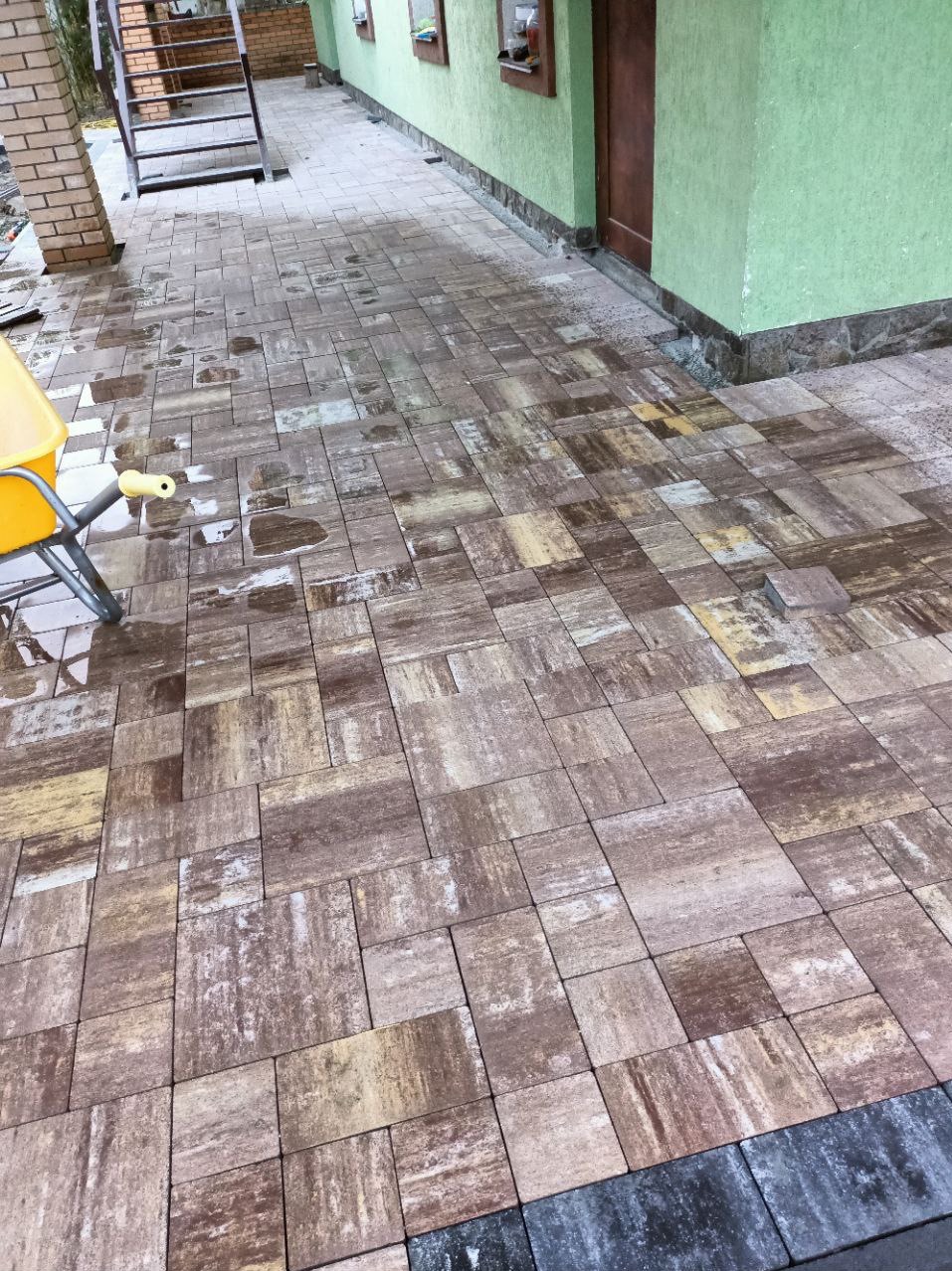 Ułożona kostka brukowa w odcieniach brązu i beżu, wzór cegiełki, fragment żółtej taczki z przodu, widoczne ślady wilgoci na powierzchni, budynek z zieloną elewacją w tle.