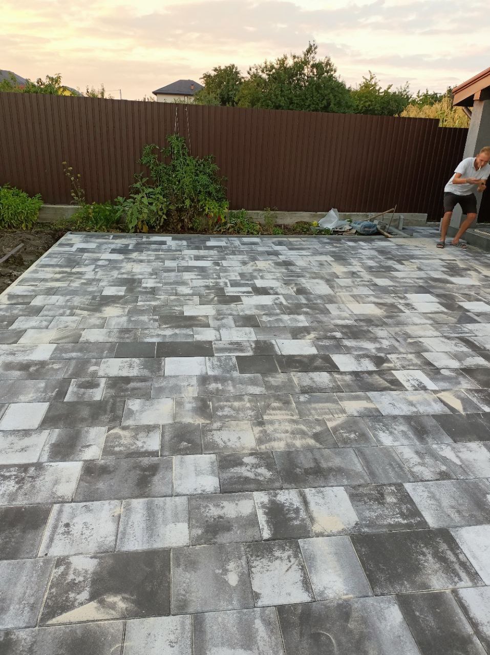 Ułożona szara kostka brukowa na posesji, widoczny fragment brązowego ogrodzenia i roślinność przy granicy z kostką, w tle sylwetka mężczyzny.