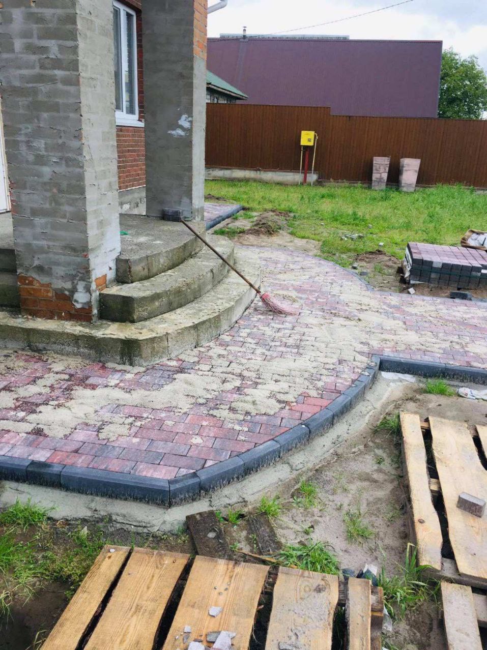 Czerwono-szara kostka brukowa ułożona w łukowaty wzór, z ciemnoszarym obrzeżem, prowadząca do betonowych schodów wejściowych, z widocznymi paletami i stertą kostki w tle.