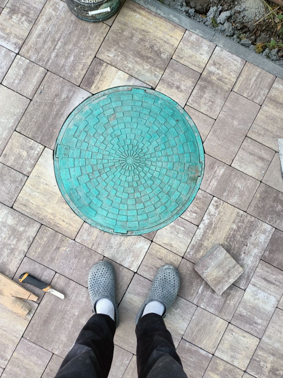 Nowo ułożona kostka brukowa w odcieniach beżu i szarości z okrągłą, turkusową pokrywą studzienki kanalizacyjnej pośrodku, widok z góry na stopy w szarych chodakach i narzędzia brukarskie.