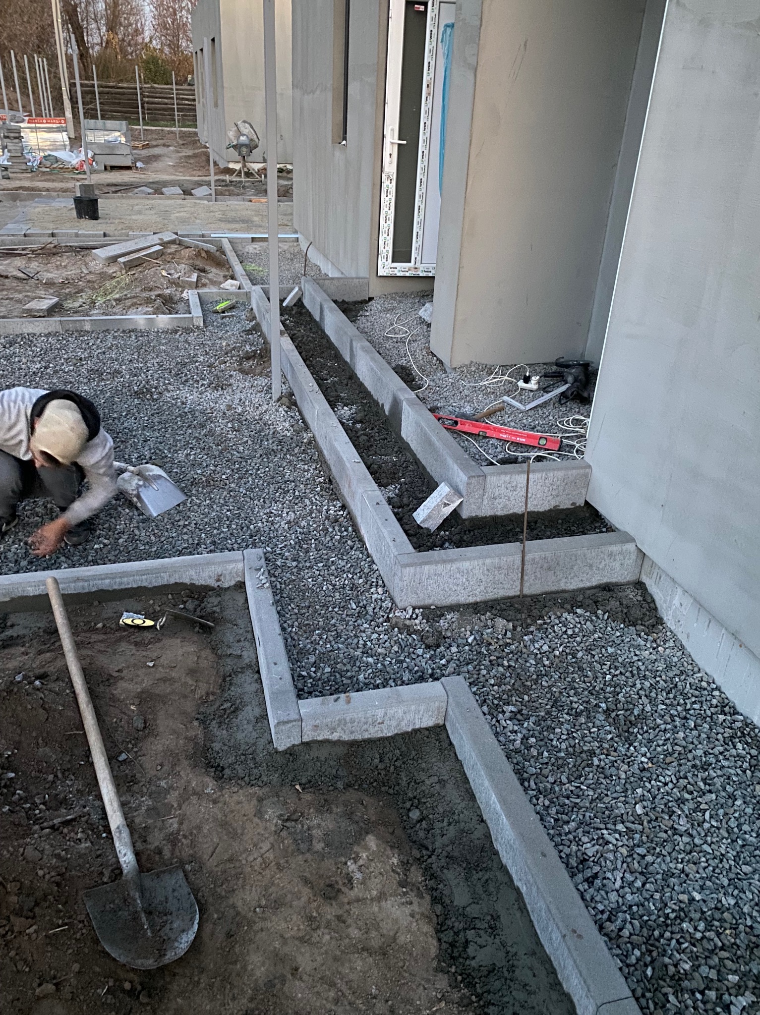 Pracownik układa obrzeża z szarej kostki betonowej wokół domu, wypełniając przestrzenie między nimi drobnym grysem; widoczna łopata i poziomica.