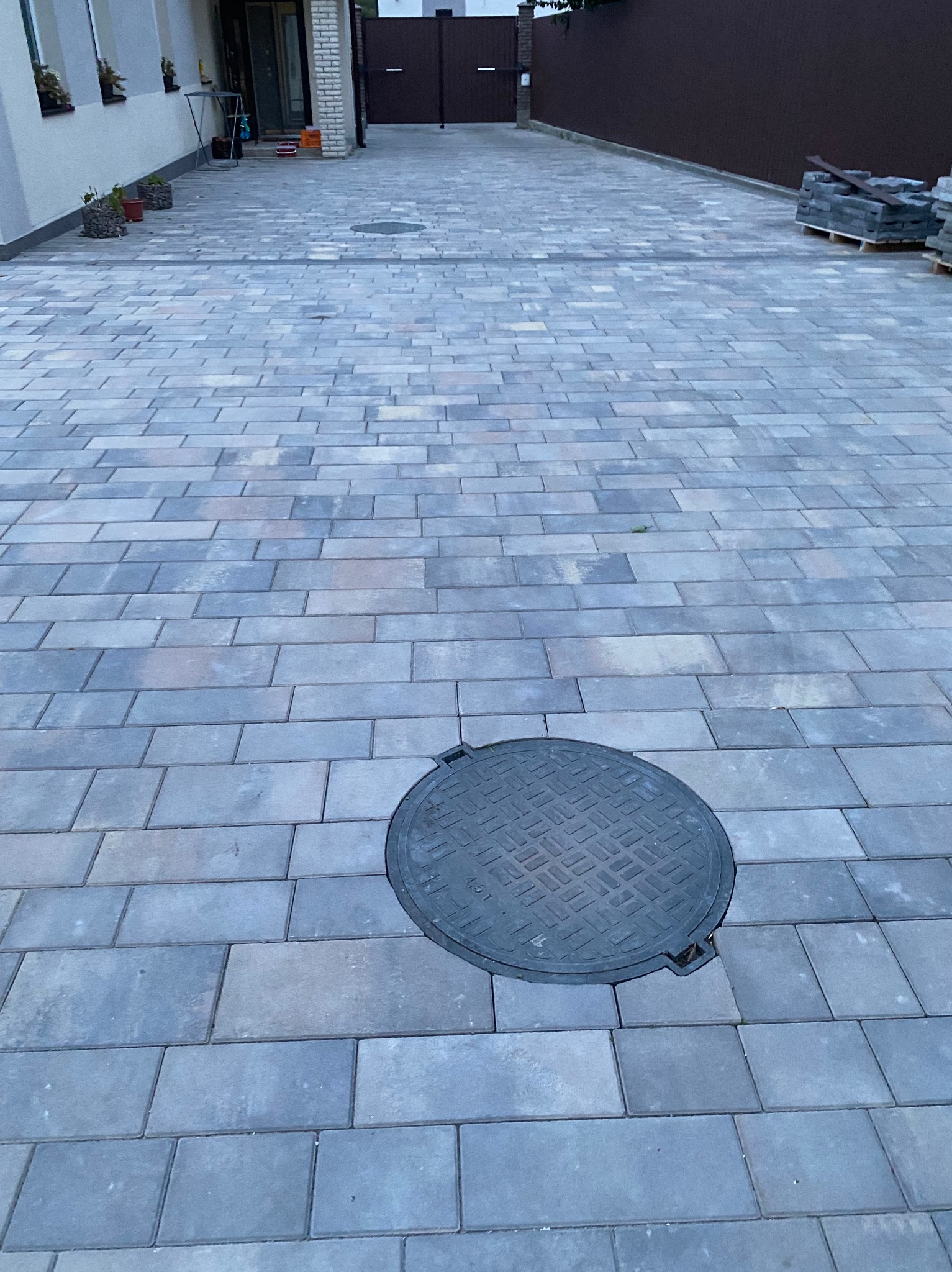 Ułożona szara kostka brukowa na podjeździe z widoczną studzienką kanalizacyjną w centrum kadru, paleta kostki brukowej w tle po prawej stronie.