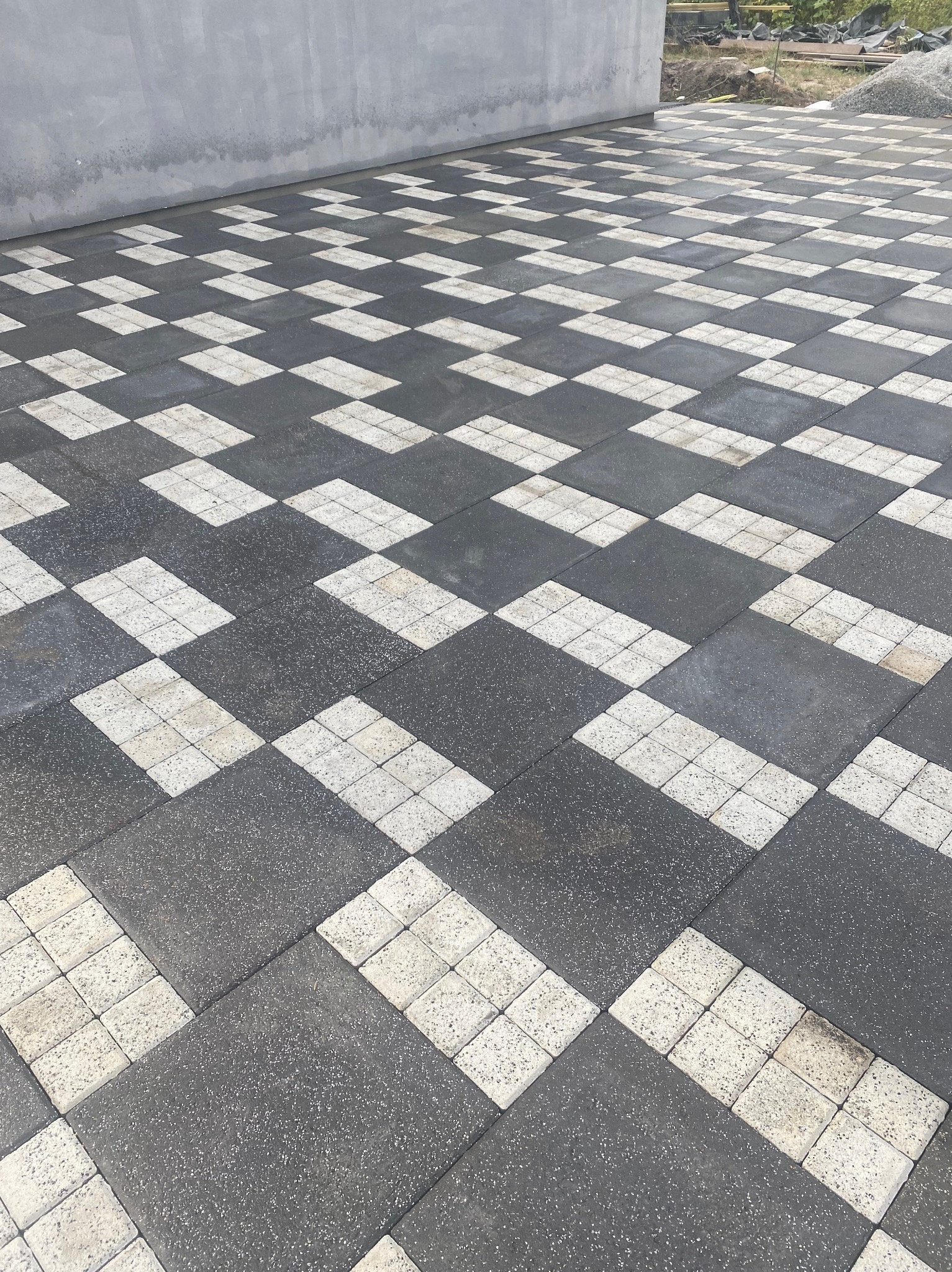 Ułożona szachownica z ciemnoszarych i jasnych kostek brukowych, widok z góry, z fragmentem szarej ściany w tle.