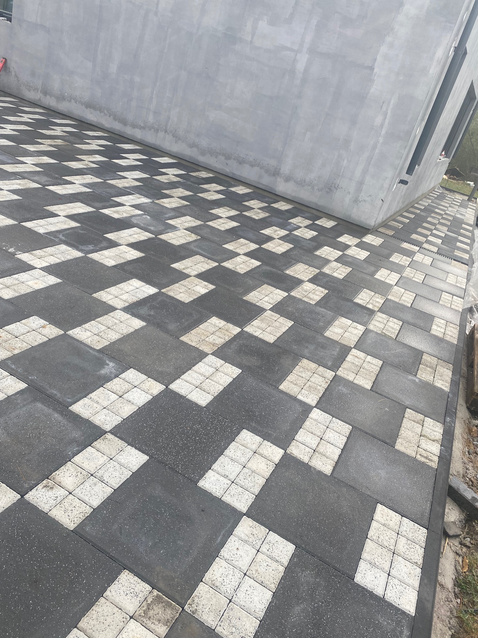 Ułożona kostka brukowa w szachownicę z ciemnoszarych i jasnych elementów przed nowoczesnym, szarym budynkiem z wąskimi oknami.