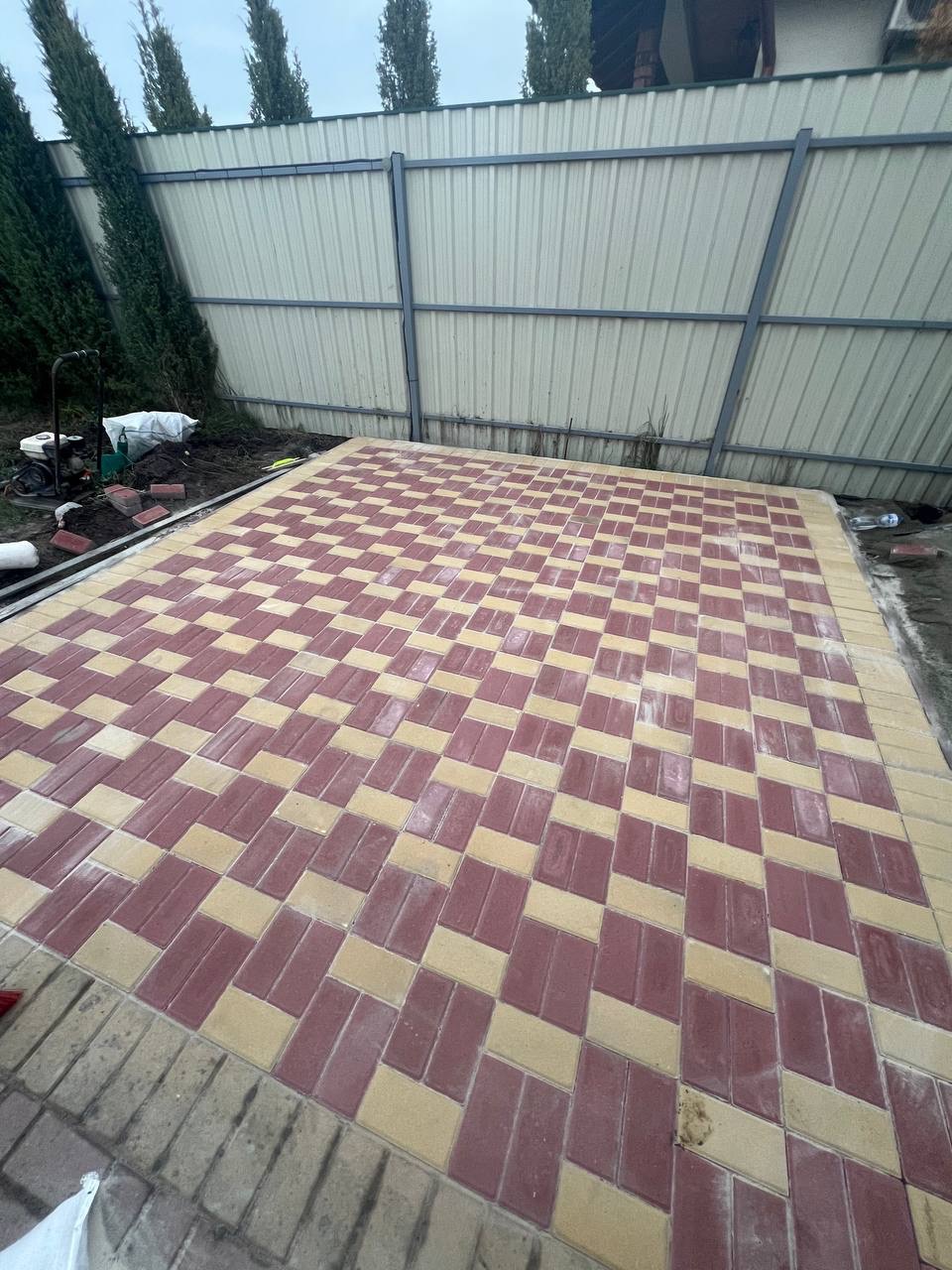 Ukończona nawierzchnia z kostki brukowej ułożonej w wzór jodełki z czerwono-beżowych prostokątnych elementów, ograniczona szarą obwódką, widoczna na tle ogrodzenia panelowego i zieleni.