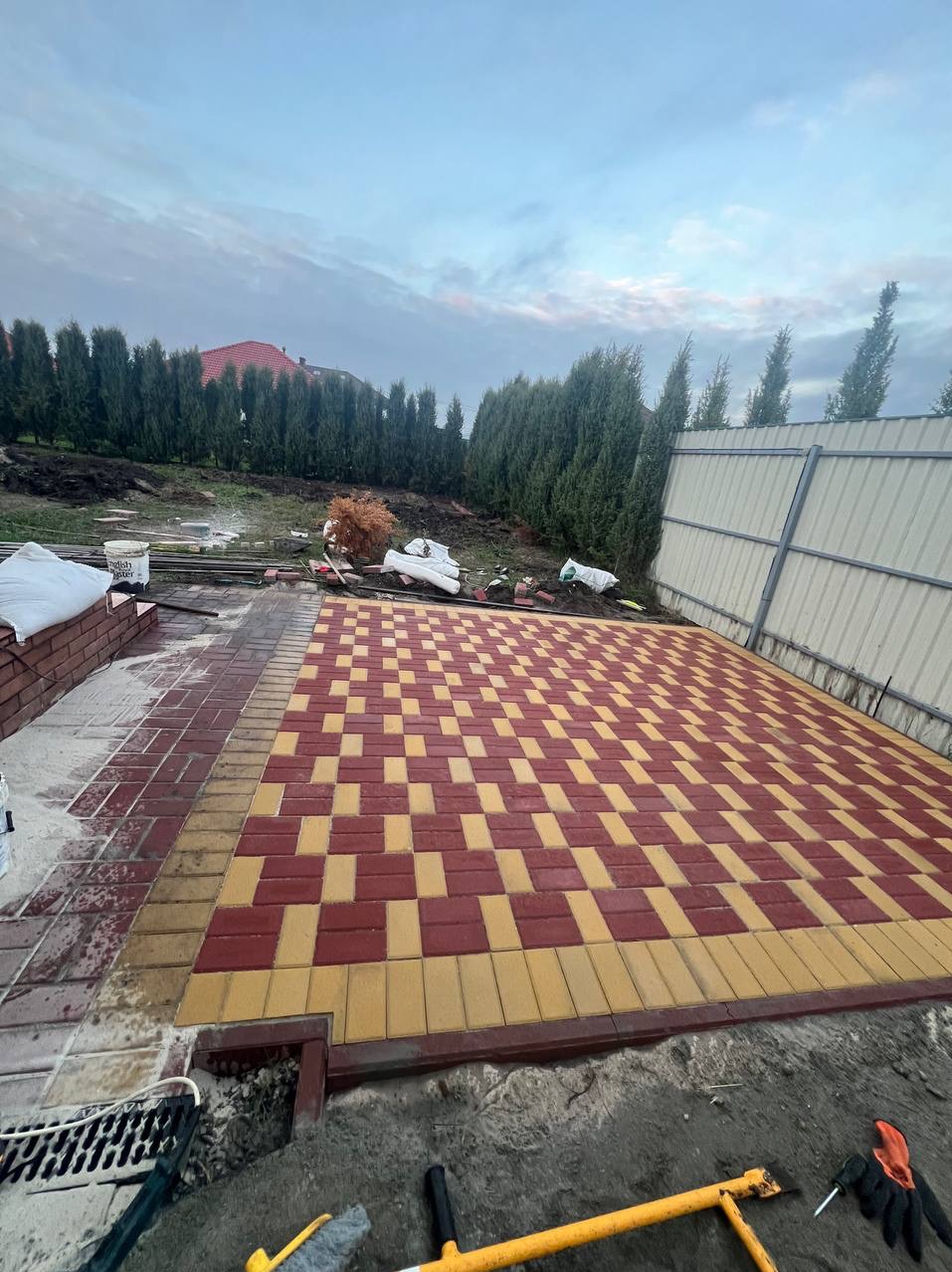 Nowa nawierzchnia z czerwono-żółtej kostki ułożona we wzór szachownicy, obrzeże z żółtej kostki, widoczne narzędzia brukarskie i fragment ogrodzenia panelowego.