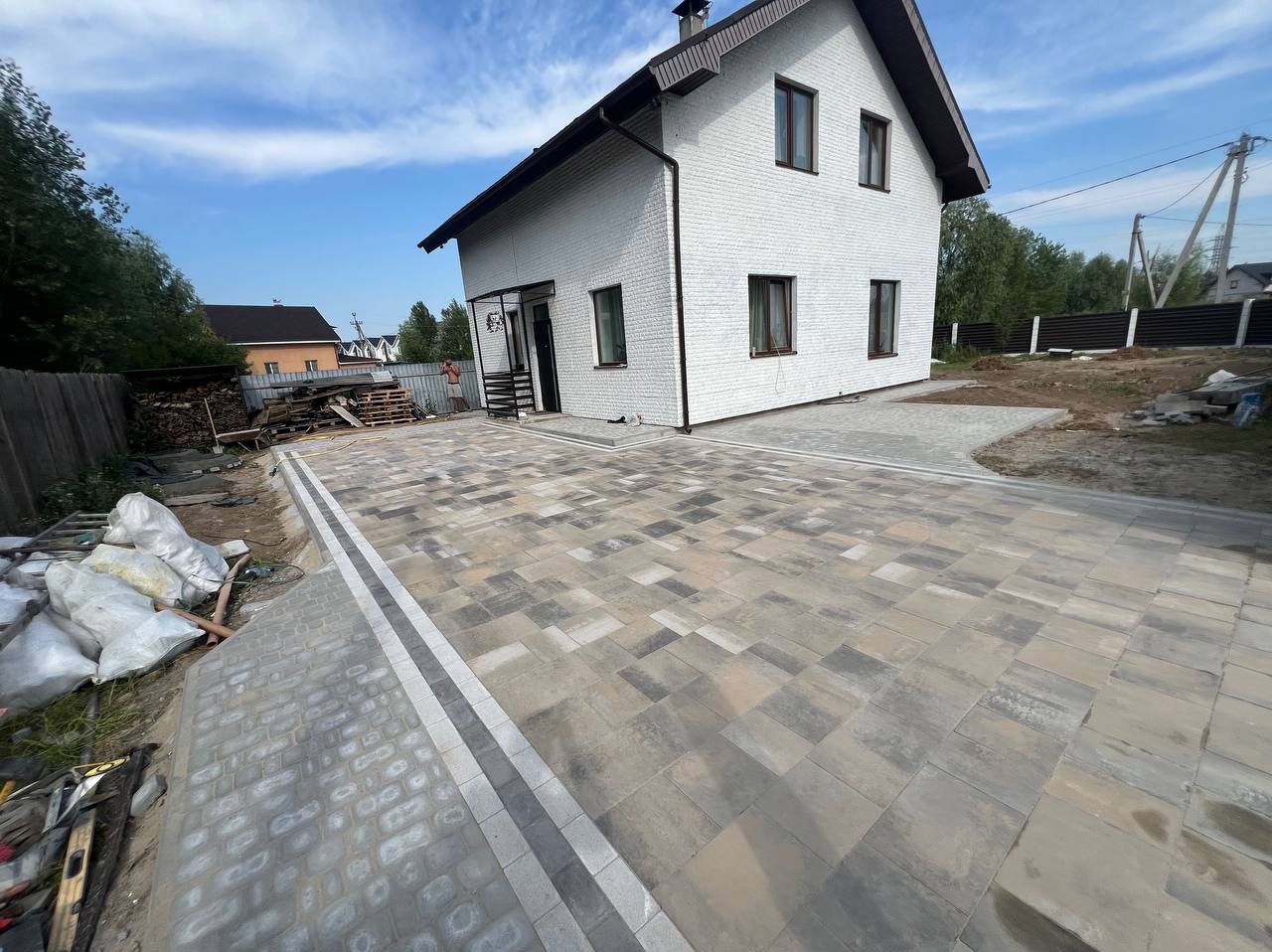 Nowoczesny podjazd z kostki brukowej w odcieniach szarości i beżu, z białymi obrzeżami, prowadzący do białego domu z cegły, widoczne worki z materiałami budowlanymi i narzędzia w tle.