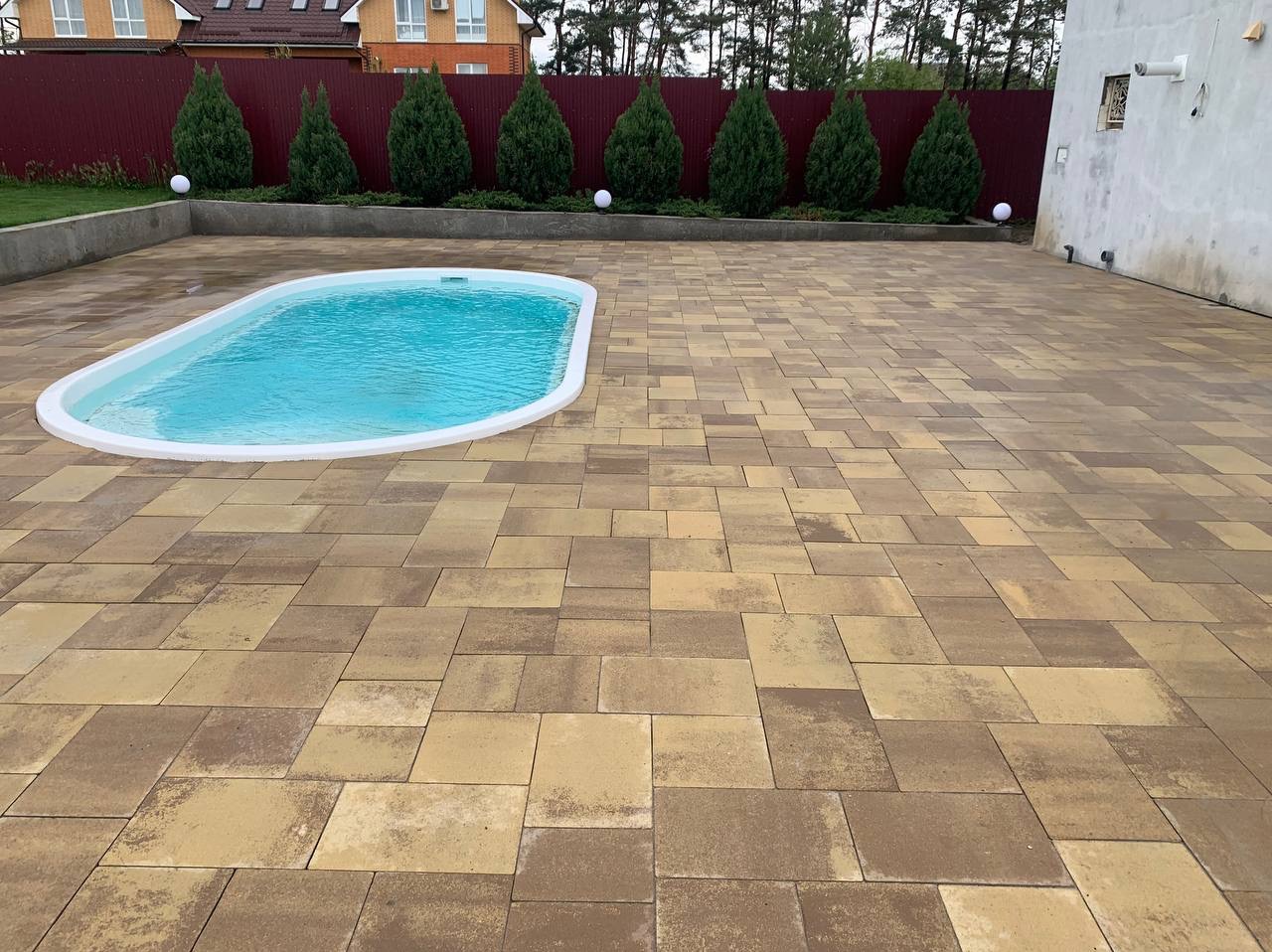 Ułożona kostka brukowa w odcieniach brązu i beżu tworzy przestrzeń wokół małego basenu ogrodowego o nieregularnym kształcie, z rzędem małych, stożkowych drzew iglastych w tle i czerwonym ogrodzeniem.